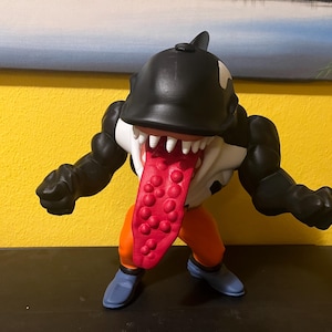 Può includere: Una statuetta di un personaggio muscoloso con una testa nera simile a un'orca, denti bianchi e una lunga lingua rossa. La figura ha braccia nere, pantaloni arancioni e scarpe blu. Lo sfondo è giallo.
