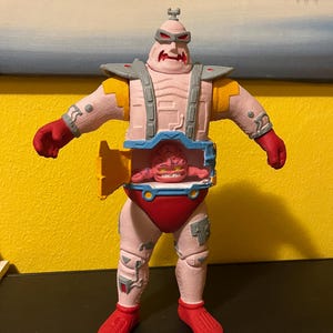 Può includere: Una action figure rosa e rossa di Krang dei Teenage Mutant Ninja Turtles. La figura ha un corpo rosa, mani e piedi rossi e una creatura simile a un cervello nella sezione centrale. Ha accenti grigi e gialli.
