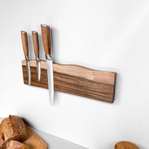 May include: A rectangular wooden magnetic knife strip with a natural live edge top, featuring a dark brown wood grain pattern. Three stainless steel kitchen knives with matching wooden handles are attached to the magnetic surface of the strip.