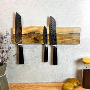 Op de afbeelding: Een magnetische messenhouder van hout met een natuurlijke houtnerf. Drie messen met zwarte handvatten en gepatenteerde messen zijn bevestigd aan de houder.
