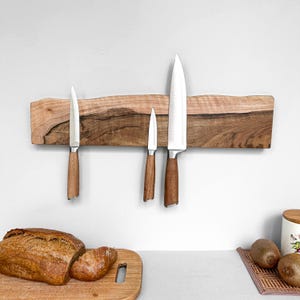 May include: A live edge wooden magnetic knife strip mounted horizontally, featuring a natural wood grain with dark and light brown tones. Three stainless steel kitchen knives with brown wooden handles are attached to the magnetic surface.