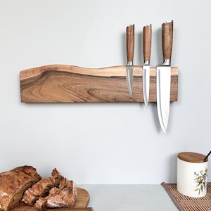 May include: A live edge wooden magnetic knife strip mounted horizontally, featuring a natural brown wood grain. Three stainless steel kitchen knives with matching wooden handles are attached to the magnetic surface in a vertical row.
