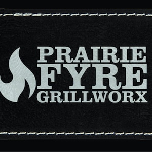 Puede incluir: Parche de cuero negro con costuras blancas alrededor de los bordes. El parche presenta un diseño de llama blanca y el texto "PRAIRIE FYRE GRILLWORX".