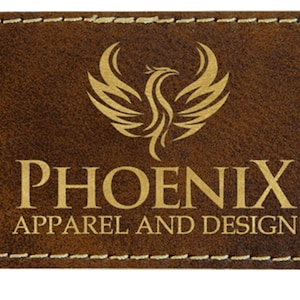 Könnte beinhalten: Rechteckiger brauner Lederaufnäher mit goldfarbenem Schriftzug "PHOENIX APPAREL AND DESIGN". Ein goldenes Phönix-Logo befindet sich über dem Text. Der Aufnäher hat einen genähten Rand.