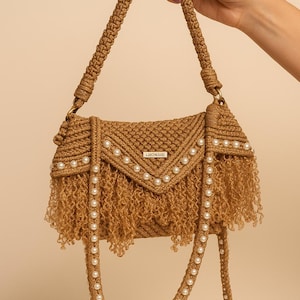 May include: A handcrafted, tan-colored macrame handbag with a shoulder strap. The bag features a fringed bottom and is adorned with rows of white pearls. The flap is decorated with pearls and a small metal tag that reads "LEHOMMAGE".