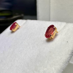 May include: A pair of gold stud earrings, each with a vibrant red oval gemstone. The earrings are set in a classic gold bezel setting, highlighting the rich colour of the stones. The studs are displayed on a white surface.