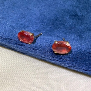Puede incluir: Un par de pendientes de botón con gemas de color rojo anaranjado, talladas en forma ovalada y engastadas en garras de metal dorado. Cada pendiente cuenta con un cierre de presión y presenta un color vibrante y translúcido.