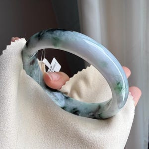 May include: A jade bangle bracelet with a smooth, polished surface. The bracelet features a blend of light green, white, and dark green hues. The jade is held by a hand on a white cloth.