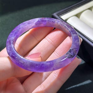 May include: A smooth, polished bangle bracelet crafted from translucent purple amethyst gemstone. The circular band features natural variations in color intensity, ranging from light lavender to deep violet, with visible internal mineral inclusions.