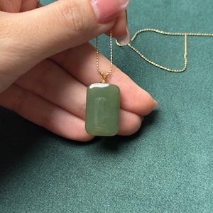 Puede incluir: Un collar de cadena dorada con un colgante rectangular de jade verde claro. El colgante es liso y pulido, con una calidad sutil y translúcida. El collar se muestra sobre un fondo verde oscuro.