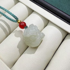 May include: A light green jade pendant carved in the shape of a mythical creature. The pendant is strung on a teal braided cord with a red bead and gold accent. The pendant is displayed on a white leatherette surface.