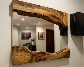 May include: Square mirror with a natural wood frame. The frame has a unique, organic shape with a light brown colour and visible wood grain. The mirror reflects a room with artwork and a door.