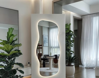 Puede incluir: Un espejo blanco moderno y de pie con una forma irregular y orgánica. El espejo está iluminado por una luz LED blanca alrededor del perímetro. El espejo refleja una habitación con paredes blancas y un techo blanco.