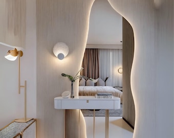 Puede incluir: Una mesa de tocador blanca con un marco de metal dorado y una encimera blanca. La mesa tiene un espejo con una forma irregular y orgánica y una lámpara en la pared detrás de ella. El espejo refleja un dormitorio con una cama y cortinas.