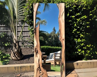 May include: A large, rectangular mirror with a natural wood frame. The frame has a unique, organic shape with knots and grain patterns. The mirror is reflecting a blue sky and green trees.