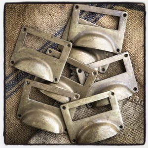 May include: A group of six antique brass drawer pulls with a rectangular cutout for a label. The pulls have a rounded, slightly domed shape and are arranged in a pile on a burlap background.