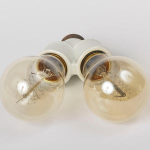 Puede incluir: Una lámpara doble de cerámica blanca con dos bombillas de vidrio transparente. Las bombillas tienen un diseño de filamento y están encendidas.