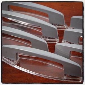 May include: A set of six gray plastic cabinet door handles with a clear plastic base. The handles are arranged in a stack, with the base of each handle visible.