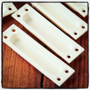 May include: Five white plastic drawer pulls with a recessed oval shape and three holes each. The pulls are arranged in a staggered pattern on a wooden surface.