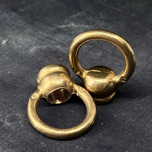 Op de afbeelding: Twee gouden metalen ringen met schroefdraad aan de uiteinden. De ringen zijn bedoeld om voorwerpen aan op te hangen.