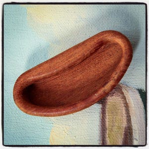 May include: A small, wooden, brown bowl with a rounded shape and a smooth, polished surface. The bowl has a slight curve in the center, creating a shallow indentation.