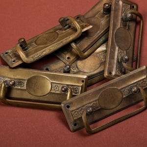 May include: A set of six antique brass drawer pulls with ornate designs. The pulls have a rectangular shape with a rounded oval in the center. The pulls are in various sizes and have a worn, aged look.