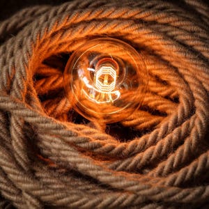 May include: A close-up of a light bulb with a warm glow, surrounded by a thick, brown rope. The rope is coiled around the bulb, creating a rustic and industrial look.