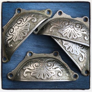 May include: A collection of antique silver-toned drawer pulls with ornate floral and scrollwork designs. Each pull has a semi-circular shape with mounting holes. The pulls are arranged on a blue wooden surface.