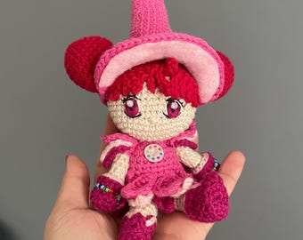 May include: A small, handmade crochet doll featuring a bright pink pointed hat, vibrant magenta hair, and large pink eyes. The doll wears a matching pink dress with a small, circular, multi-colored button at the center and has small beaded bracelets on its wrists.