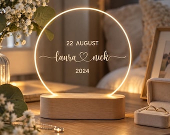 Puede incluir: Un letrero luminoso LED circular con base de madera, grabado con el texto 22 AUGUST, laura, un símbolo de corazón, nick y 2024. La lámpara emite una luz cálida y se encuentra junto a un pequeño joyero con dos anillos de oro.