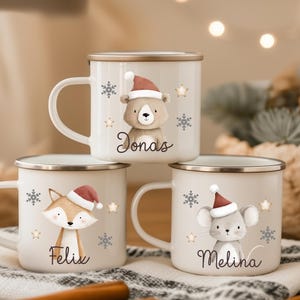 Könnte beinhalten: Drei weiße Emaille-Tassen mit festlichen Tiermotiven. Jede Tasse zeigt ein anderes Tier mit Weihnachtsmütze, mit den Namen "Jonas", "Felix" und "Melina" darunter. Schneeflocken und Sterne verzieren die Tassen.