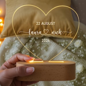 以下が含まれることがあります： 「Laura & Nick」と「22 August 2024」の日付が刻まれたハート型のLEDアクリルランプ。木製の台座に置かれ、温かい光を放ちます。結婚式の装飾に最適です。