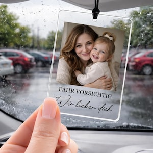 Può includere: Un profumatore per auto rettangolare in acrilico trasparente con la foto di una donna e un bambino. Il profumatore ha il testo tedesco "FAHR VORSICHTIG Wir lieben dich" e pende da un cordino nero. Lo sfondo mostra un parcheggio piovoso.
