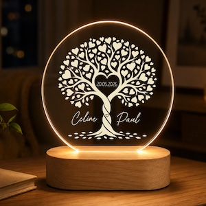 Könnte beinhalten: Ein personalisiertes LED-Nachtlicht mit einem Baumdesign und herzförmigen Blättern. Die transparente Acrylplatte zeigt die Namen "Celine" und "Paul" mit einem Datum, auf einem Holzsockel. Das Licht strahlt ein warmes Leuchten aus.