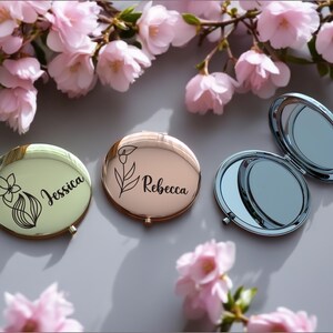 Puede incluir: Tres espejos compactos personalizados. Uno está abierto, revelando una superficie reflectante. Los otros dos están cerrados, uno con el nombre "Jessica" y un diseño floral, el otro con "Rebecca" y un diseño floral. Flores rosas en el fondo.