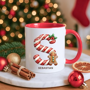 Könnte beinhalten: Weißer Keramikbecher mit rotem Innenraum und Griff. Der Becher zeigt ein Zuckerstangen-Design des Buchstabens "S" mit einem Lebkuchenmann und dem Namen "Sebastian". Festlicher Weihnachtsbecher mit Weihnachtsdekorationen.