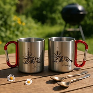 May include: Two stainless steel camping mugs with red carabiner handles sit on a wooden table. Each mug features a compass and mountain design with a name. Two gold spoons and daisies are also on the table.