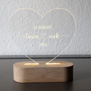 May include: An illuminated heart-shaped acrylic light, engraved with the names "Laura & Nick" and the date "22 August 2024". It rests on a rectangular wooden base, emitting a warm light.