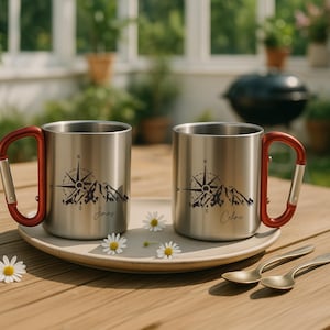 May include: Two stainless steel camping mugs with red carabiner handles sit on a plate with daisies. Each mug features a compass and mountain design with a name. Two small gold spoons are next to the plate.