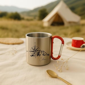 May include: A stainless steel camping mug with a red carabiner handle. The mug features a black mountain and compass design with the name "Celine" in script. A small red cup and spoon are in the background.