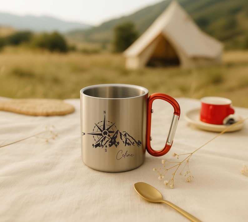 May include: A stainless steel camping mug with a red carabiner handle. The mug features a black mountain and compass design with the name "Celine" in script. A small red cup and spoon are in the background.
