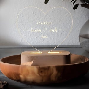 May include: An illuminated acrylic heart wedding decoration, engraved with "Laura & Nick" and the date "22 August 2024". The light sits on a wooden base, inside a wooden bowl. A modern, romantic keepsake for a wedding or anniversary.