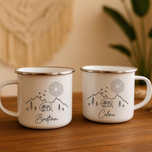 May include: Two white enamel camping mugs with black line art of mountains, a sun, trees, and a camper. One mug has the name "Bastian" and the other has "Celine" in cursive. Both mugs have a silver rim.