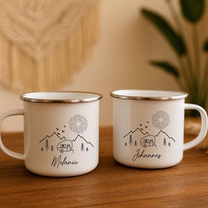 May include: Two white enamel mugs with silver rims, each featuring a black mountain and camper design. One mug has the name "Melanie", the other "Johannes". The design includes a sun, birds, and trees.