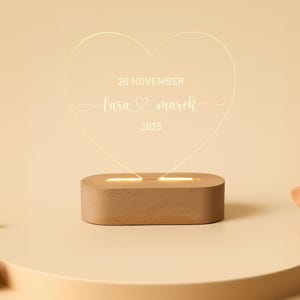 May include: A heart-shaped LED light with the text "20 NOVEMBER, Lara & Marek, 2025" displayed on a wooden base. The light emits a warm glow, ideal for a wedding or anniversary gift. The background is a soft beige.