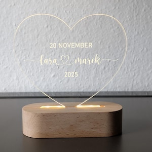 May include: An illuminated heart-shaped acrylic sign, engraved with the names "Lara & Marek" and the date "20 November 2025". The clear acrylic heart sits on a rectangular wooden base, providing a warm, ambient light.