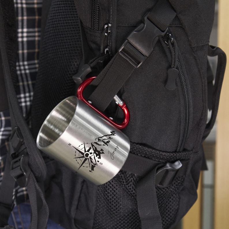 May include: A stainless steel camping mug with a red carabiner clip attached to a black backpack. The mug features a black compass rose design with a mountain scene and the name "Jones" in script.