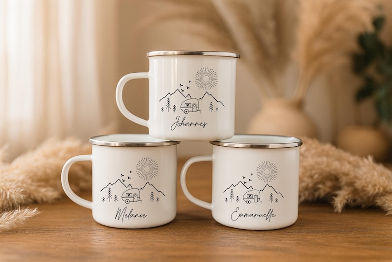 May include: Three white enamel mugs with silver rims are stacked on a wooden surface. Each mug features a black line drawing of a mountain scene with a camper, sun, and personalized name. The names are Johannes, Melanie, and Emmanuelle.