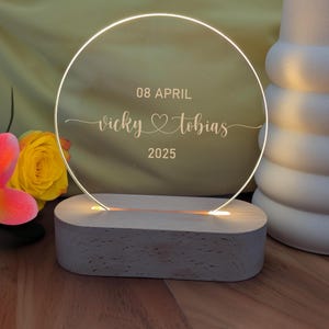 May include: A circular illuminated acrylic wedding decoration, engraved with "Vicky & Tobias" and the date "08 APRIL 2025". The clear acrylic is lit by a warm light and rests on a wooden base. A yellow rose and a pink and orange flower are in the background.