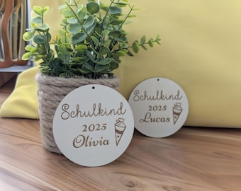 May include: Two round wooden ornaments with the text "Schulkind 2025" and the names "Olivia" and "Lucas" written on them. Each ornament has a small illustration of a school bag.